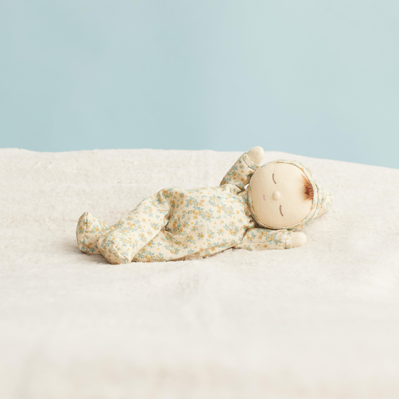 A small doll or stuffed baby figure wearing a cream pajama set with a light blue and yellow floral pattern, lying on its back on a soft white blanket against a pale blue background.
