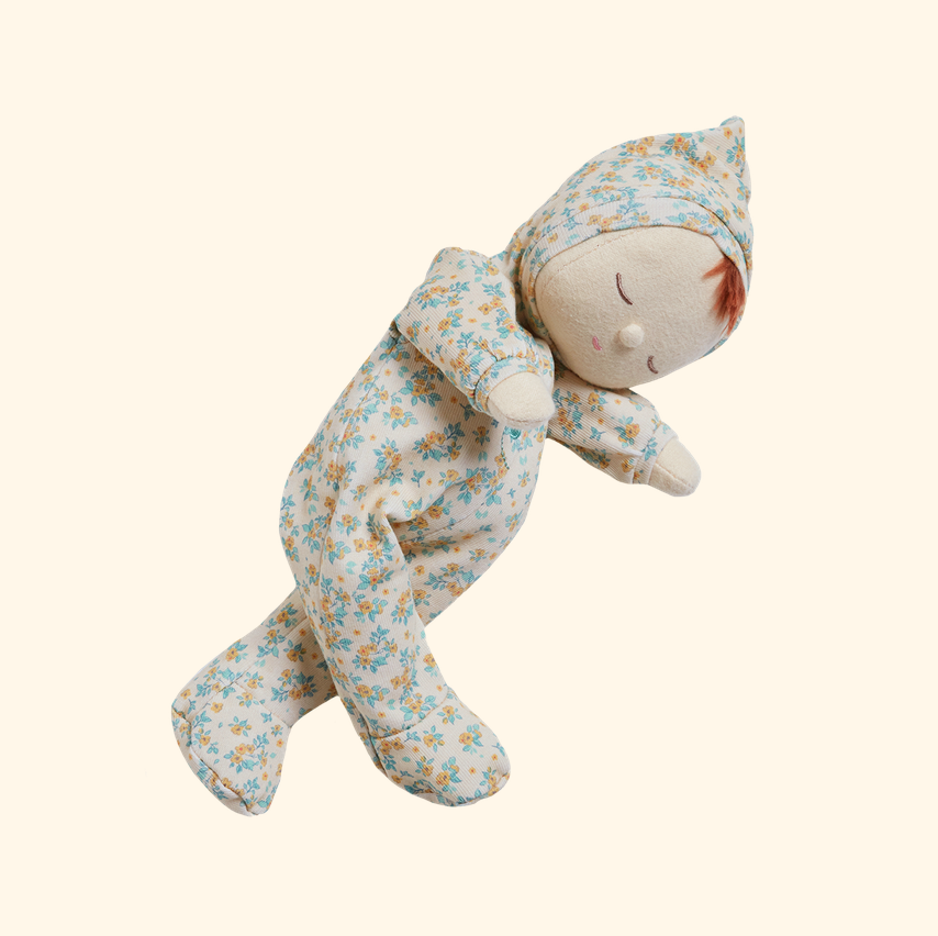 A plush baby doll wearing a light cream footed onesie with a blue and yellow floral print, posed as if sleeping on a solid pale beige background.