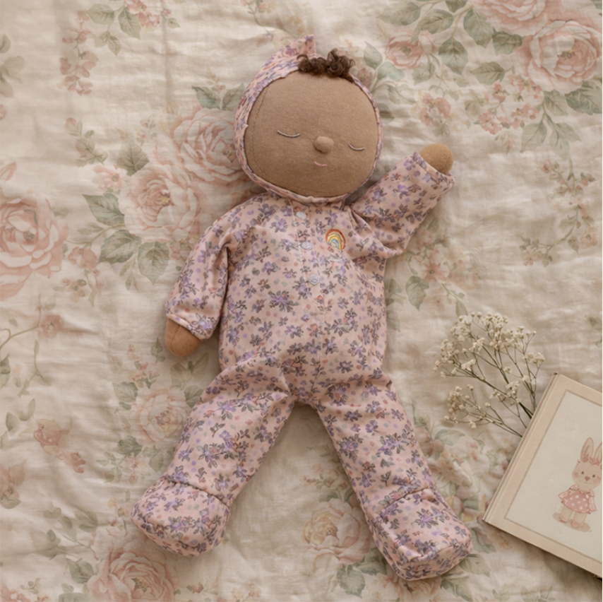 A plush baby doll wearing a pink floral footed onesie and bonnet, lying on a matching floral blanket with dried flowers and a small framed bunny picture nearby.