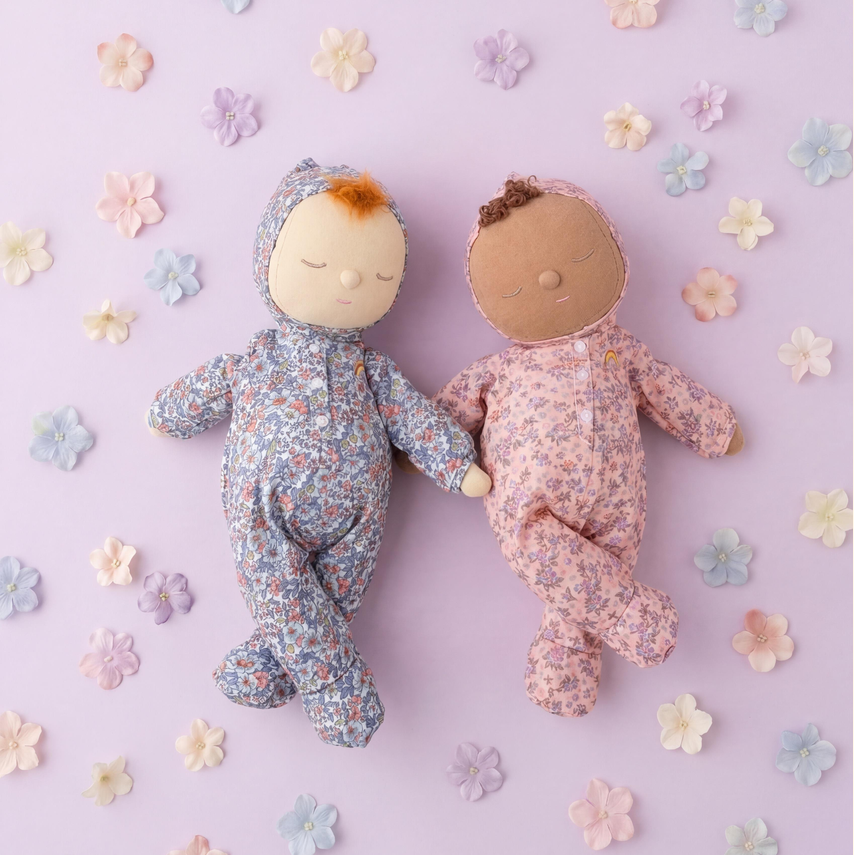 Two handmade fabric baby dolls lying side by side on a pastel lavender background decorated with small paper flowers. The doll on the left wears a blue floral onesie and has light skin with orange hair, while the doll on the right wears a pink floral onesie and has medium skin tone.