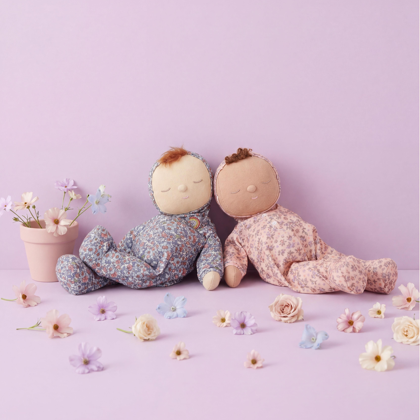 Two baby doll figures lying side by side on a pastel purple surface, one wearing a blue floral onesie and the other a pink floral onesie, with small flowers and a pink flower pot around them.
