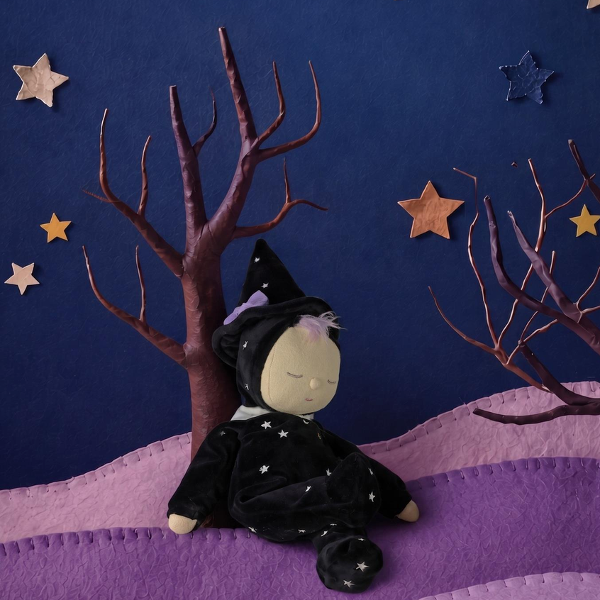 A small witch doll wearing a black pointed hat and black dress with white star details, sitting on a purple felt landscape with bare brown trees and star decorations against a dark blue background.