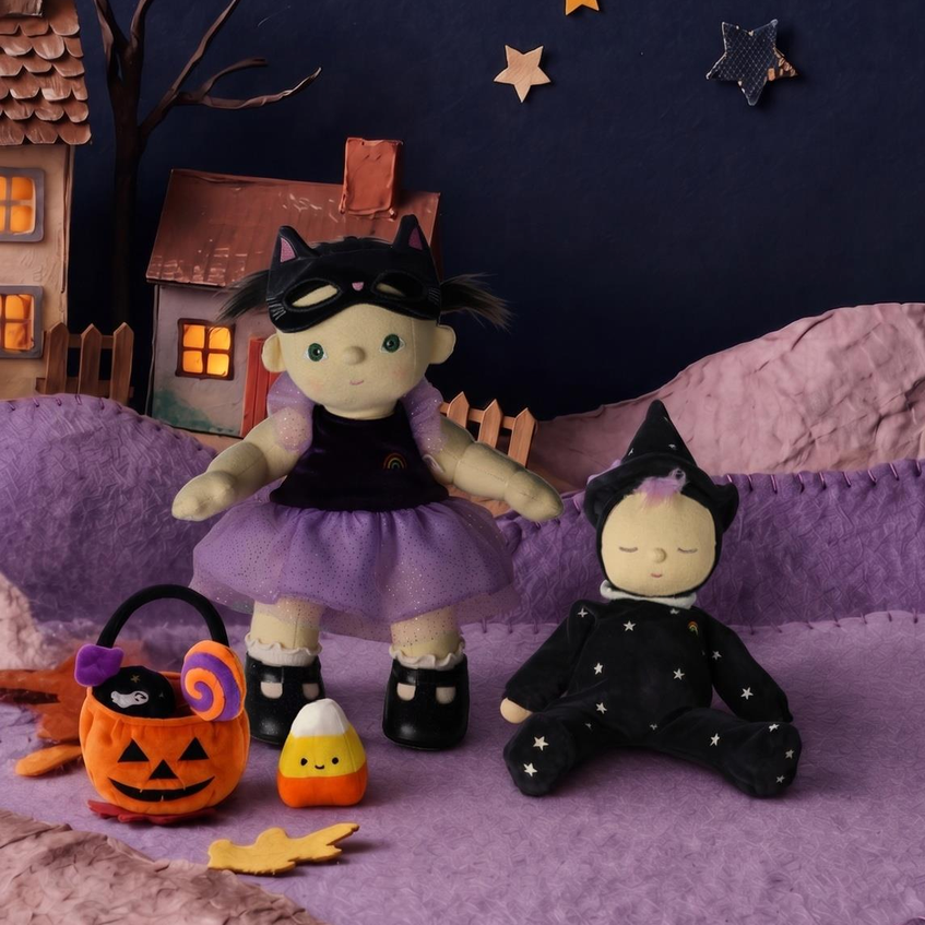 A Halloween-themed doll display featuring a witch doll in a purple dress and a baby doll in a black outfit, styled with a jack-o'-lantern bucket, candy corn, and a small house against a dark blue starry backdrop.