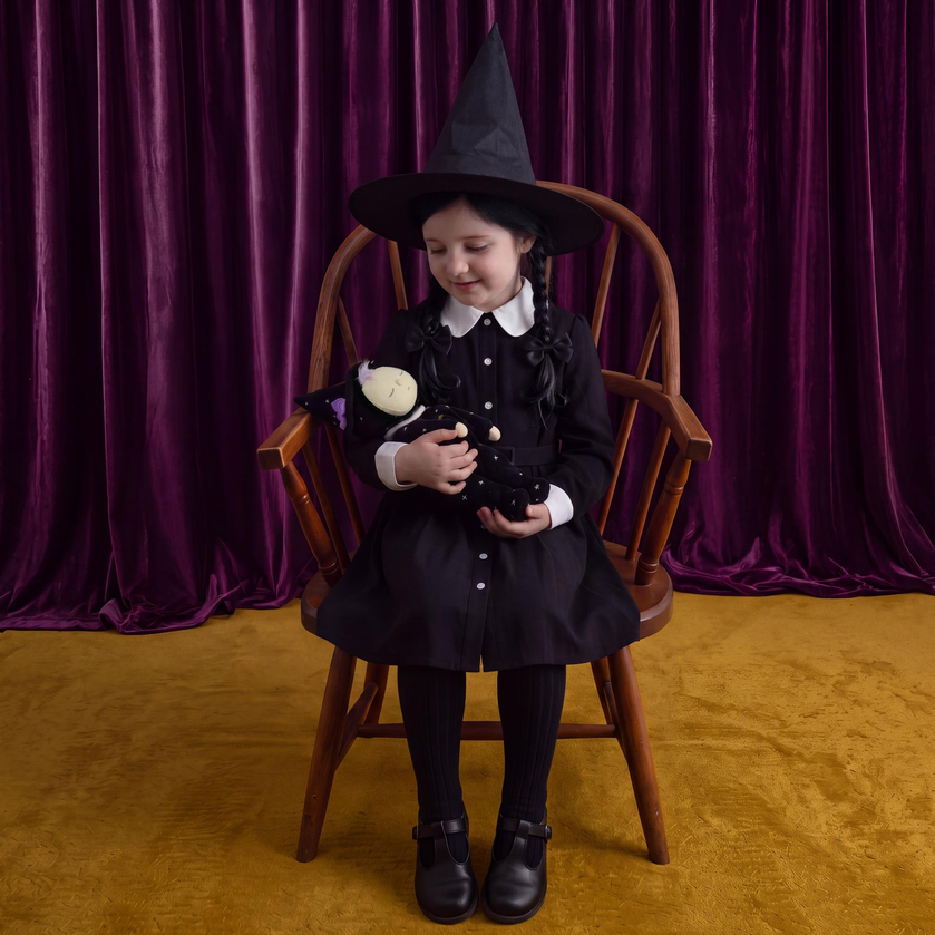 A young girl dressed as a witch sits in a wooden chair against a purple velvet curtain backdrop. She wears a black witch hat, a black dress with a white collar, black tights, and black shoes, while holding a small black and white plush doll.