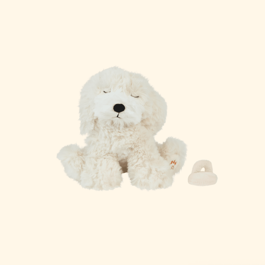 A fluffy white plush dog toy with closed eyes and a small matching white ring-shaped accessory beside it, photographed on a plain warm cream background.