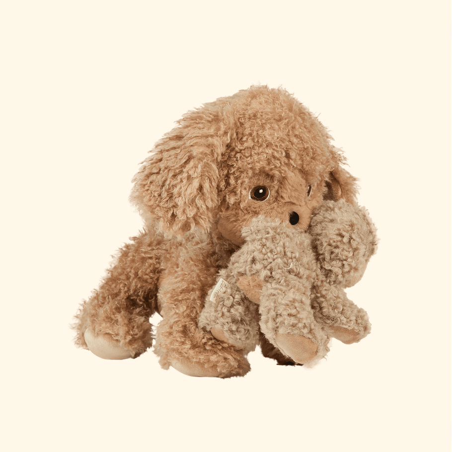 A plush stuffed animal shaped like a curly-haired dog sitting and holding a small stuffed figure in its paws, shown on a plain warm off-white background.