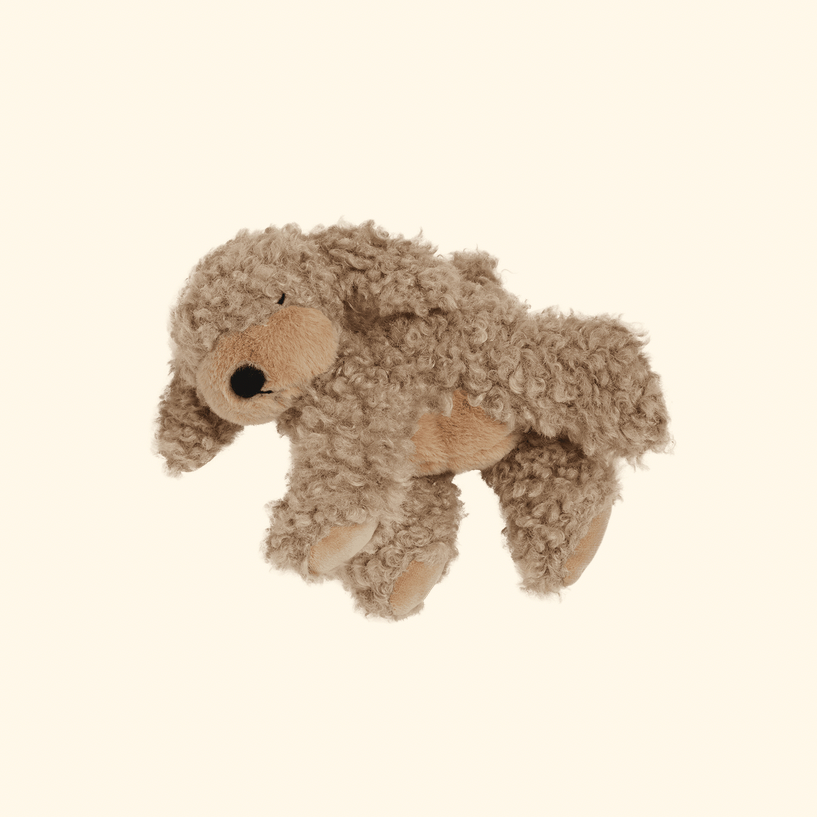 A small plush stuffed animal shaped like a dog or bear, made of curly brown faux fur with a tan face, belly, and paws, photographed on a plain warm cream background.