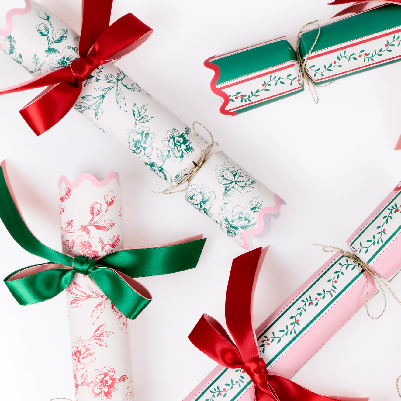 Three Christmas-themed paper cracker ornaments and wrapped gift boxes arranged on a white background, decorated with floral patterns in green and pink, tied with red and green satin ribbons.