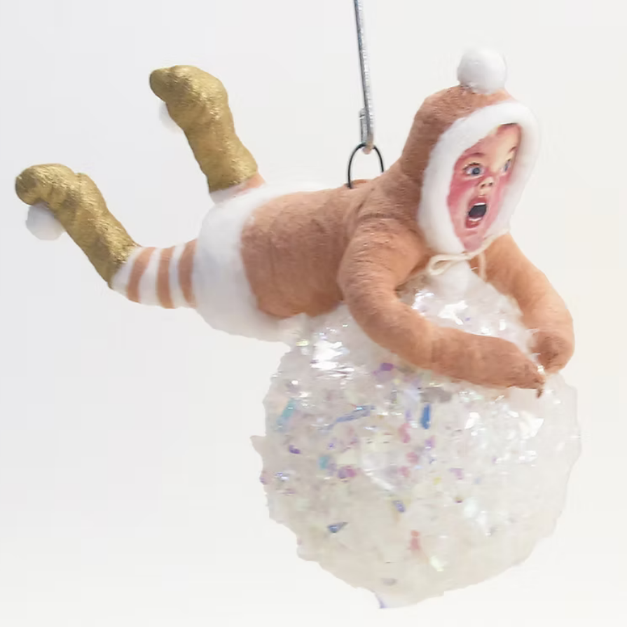 A novelty hanging ornament shaped like a baby in a brown hooded outfit with gold glittery feet, white diaper details, and a printed face, posed above a translucent glittery white ball on a plain white background.