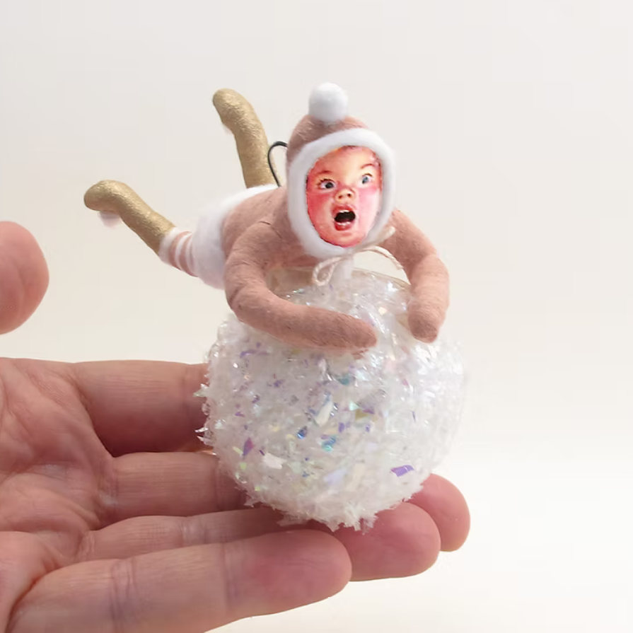 A miniature plush ornament of a baby figure wearing a brown and white onesie, sitting inside a white glittery ball, held in a person's hand against a plain white background.