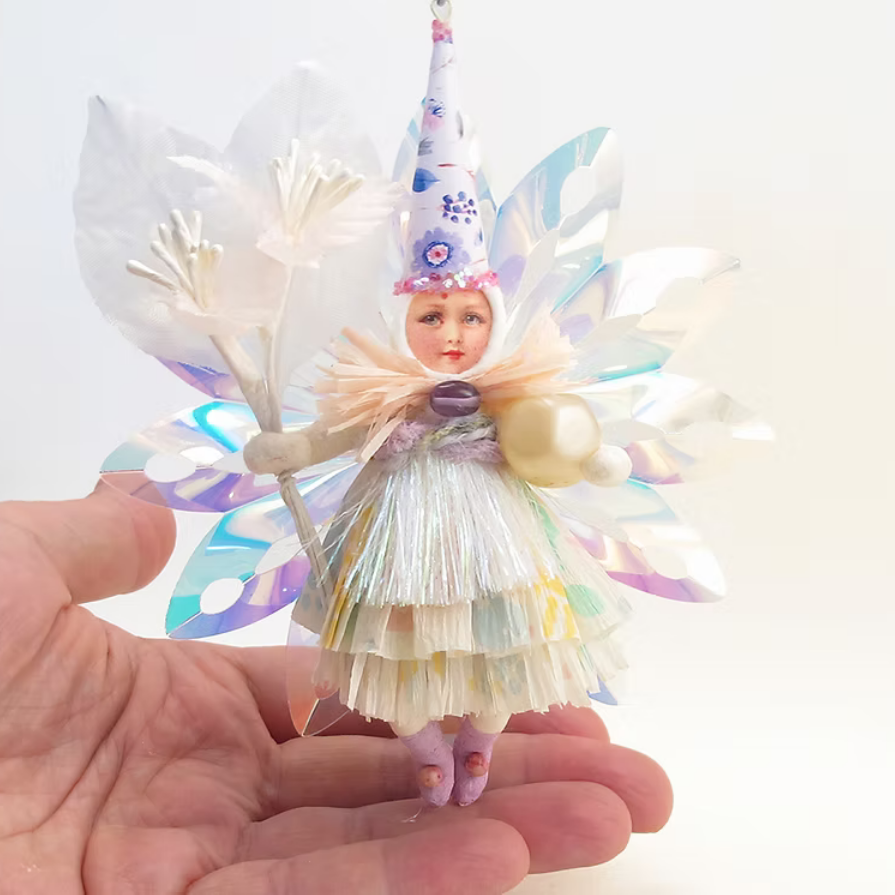 A handmade fairy ornament held in a person's hand against a plain white background. The ornament features a small doll with a pointed hat, iridescent butterfly wings, and a layered ruffled dress in pastel colors.