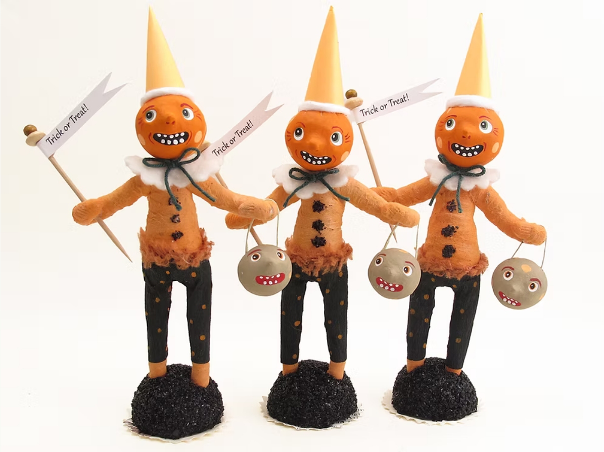 Three orange gingerbread-style trick or treat figurines with yellow pointed hats, white ruffled collars, black polka-dot pants, and black glittery bases, each holding a small gray pumpkin and a sign that reads 'Treat or Trick!'.
