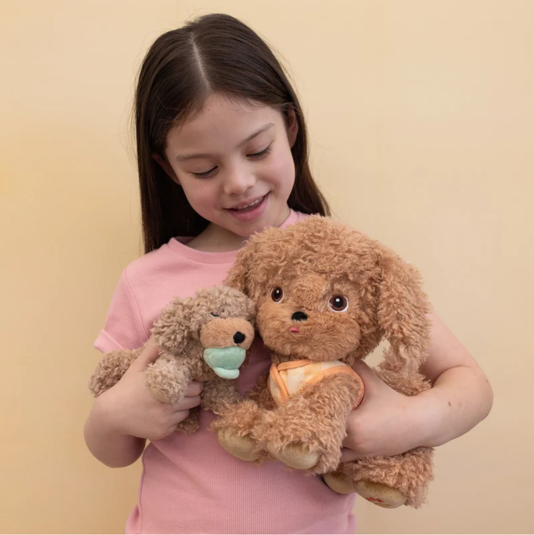 A young girl with long dark hair, wearing a pink t-shirt, is smiling and holding two plush toy dogs against a solid beige background.