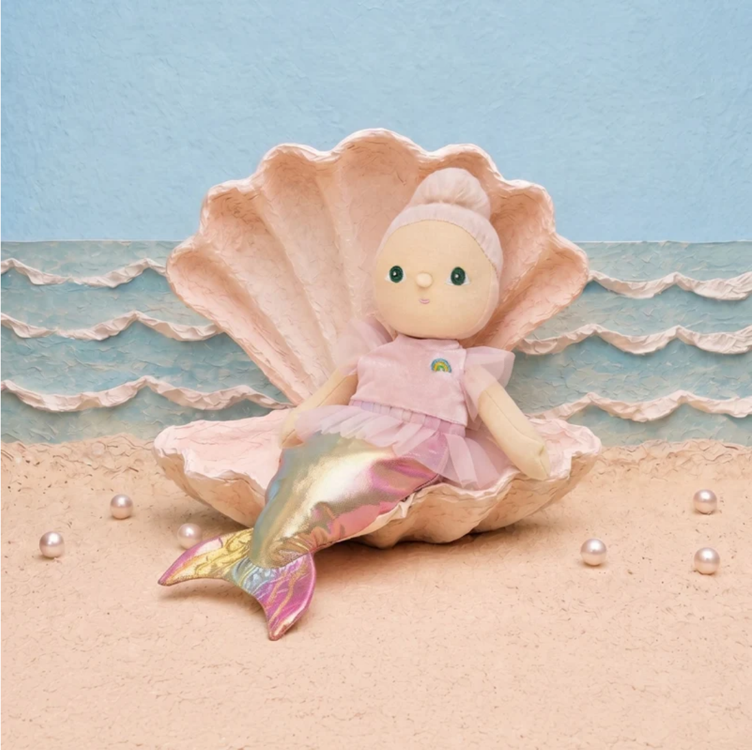 A mermaid doll with a pink dress and iridescent tail sits inside a large open seashell on a sandy beach set with blue ocean waves and pearl-like beads.