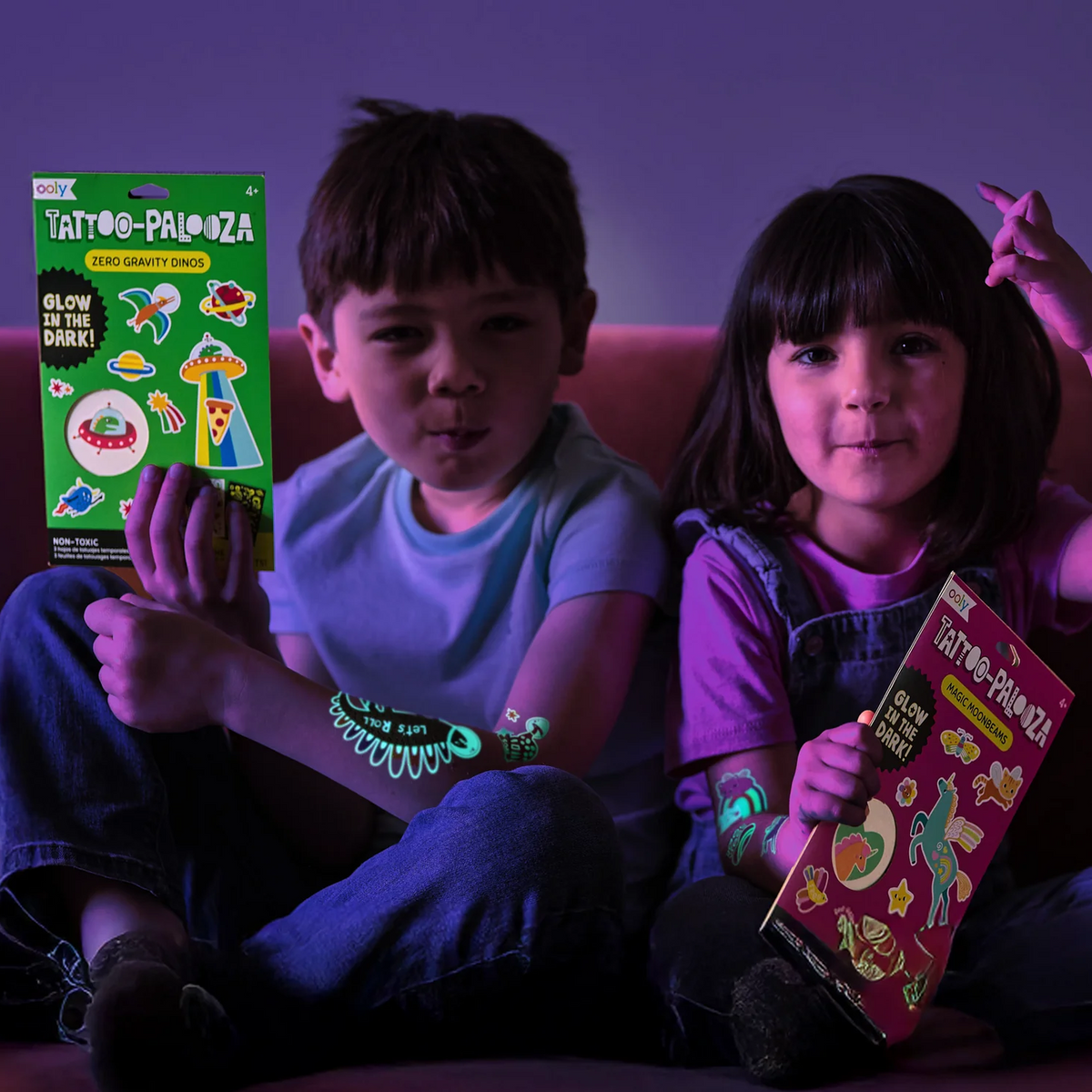 Two children sitting on a couch in dim purple lighting, each holding a boxed glow-in-the-dark tattoo palette product. The boxes are labeled 'TATTOO-PALOZA' and feature colorful illustrated designs.