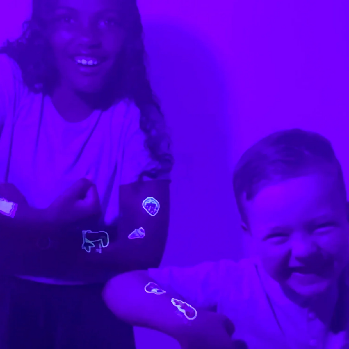Two children smiling under a purple UV light, wearing glow-in-the-dark temporary tattoos on their arms.