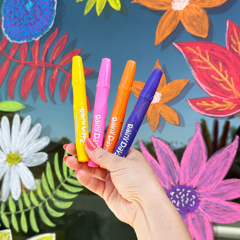 A hand holds four colorful Rain Day highlighters in yellow, pink, orange, and purple against a vibrant, hand-painted floral background.