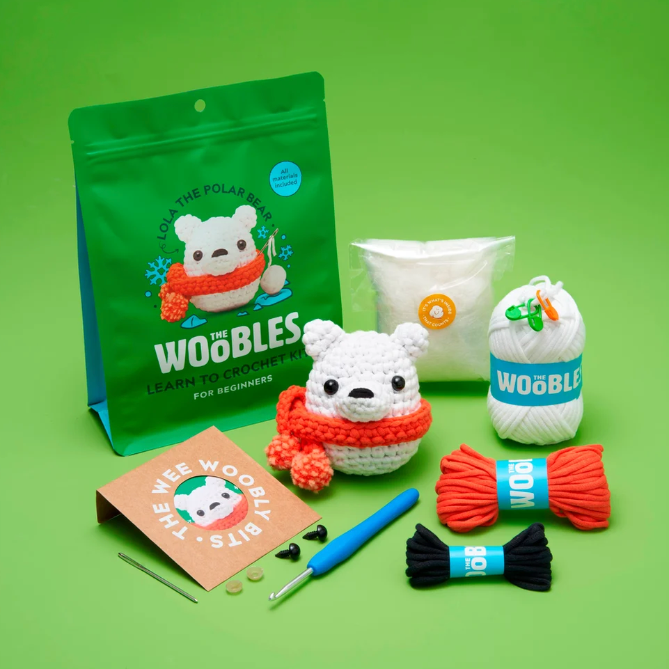 A crocheted kit for designers by WooBles The Bear Pips, featuring a completed white polar bear crochet doll with an orange-red scarf, alongside the green packaging pouch, white yarn, orange yarn, black yarn, a blue crochet hook, a metal needle, and a small instruction card on a solid green background.