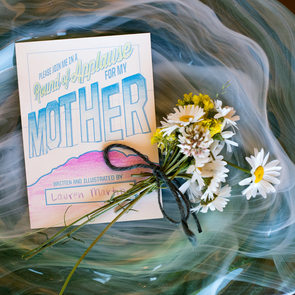 Book titled 'Please Join Me in a Round of Applause for My Mother' with a bouquet of flowers on a textured surface.