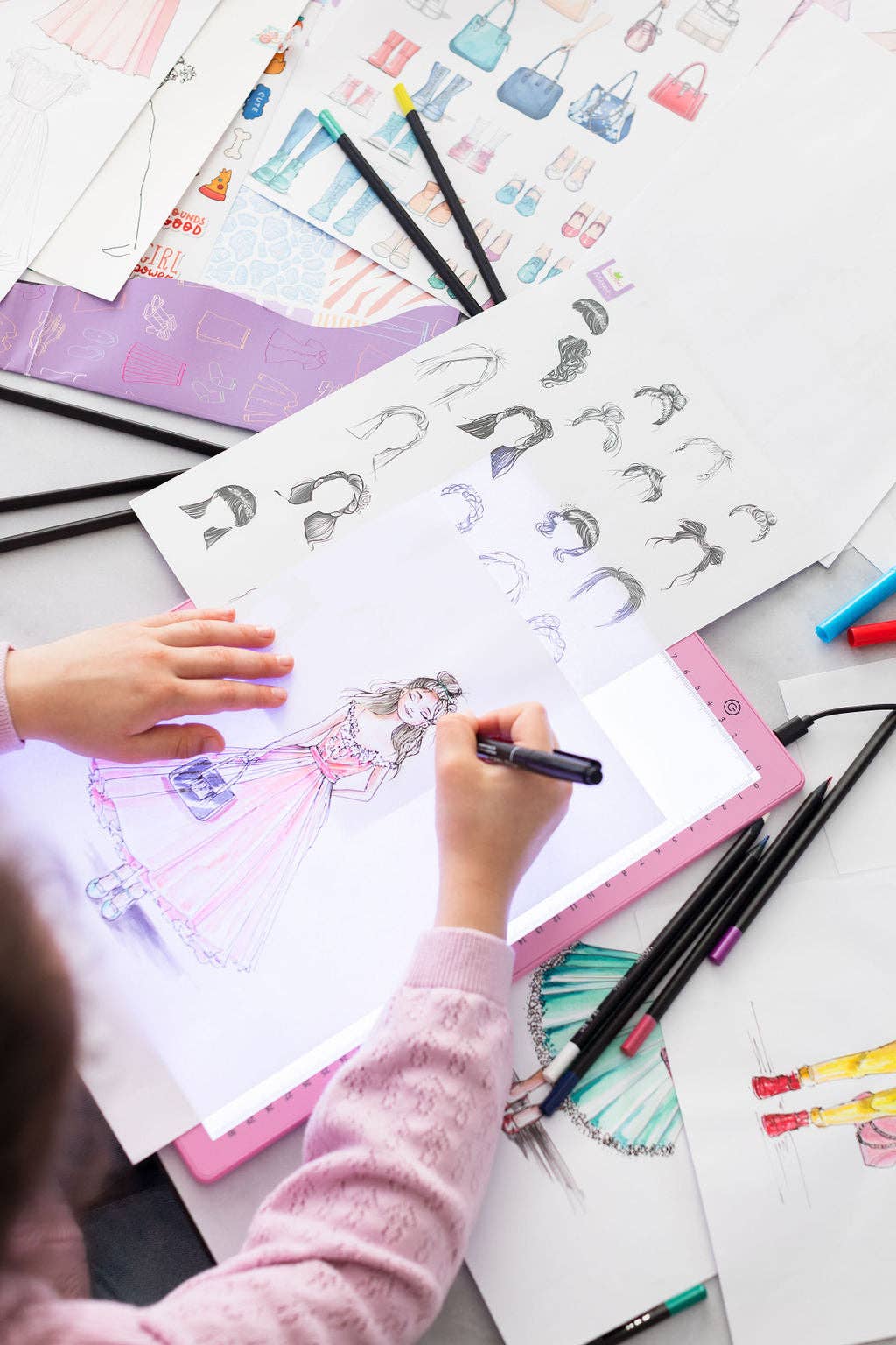 Child drawing fashion illustrations with markers on a table.