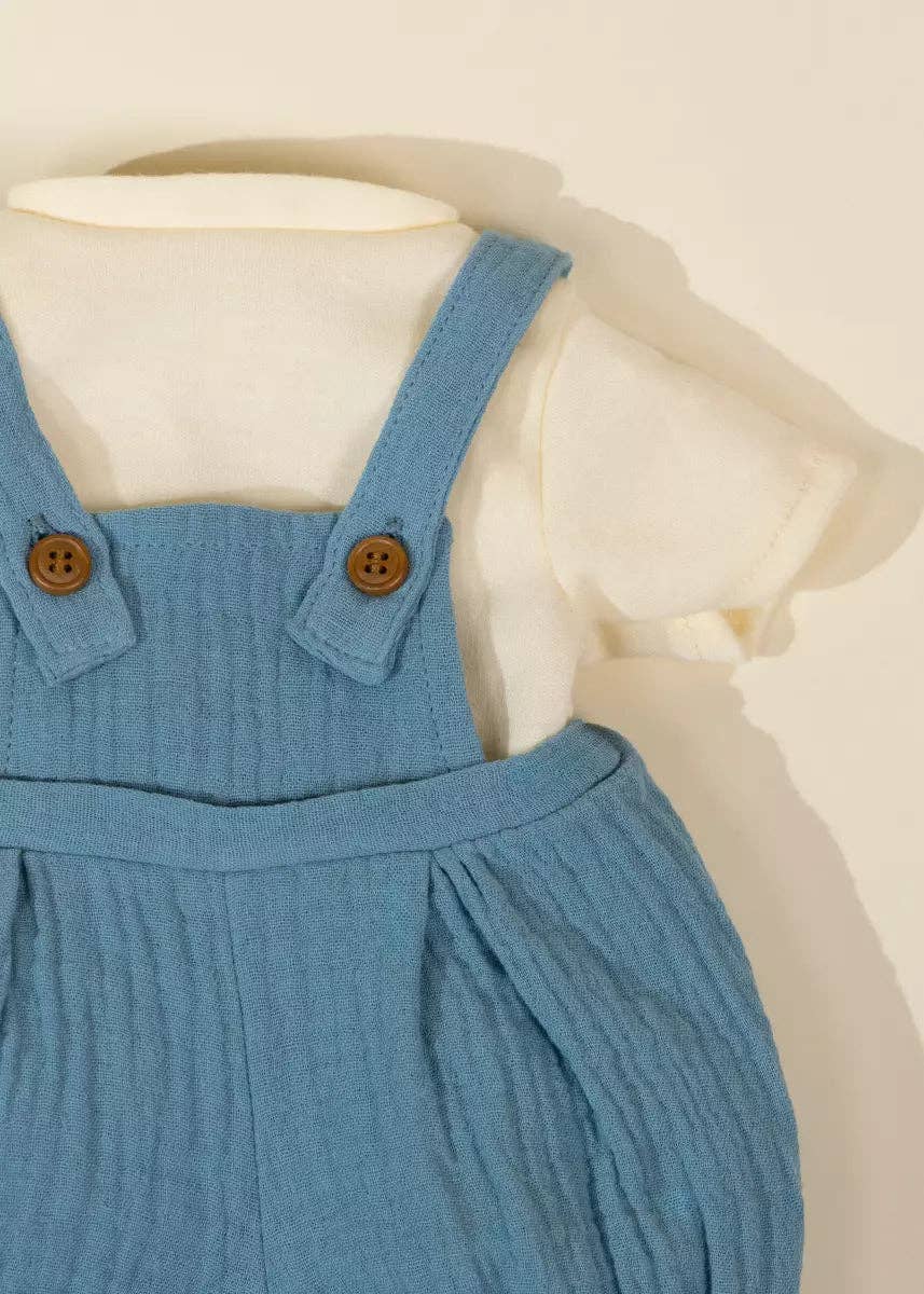 A close-up of a baby outfit featuring a light blue ribbed corduroy-style overall dress with brown buttons, layered over a short-sleeve cream shirt, displayed on a cream mannequin against a warm beige background.