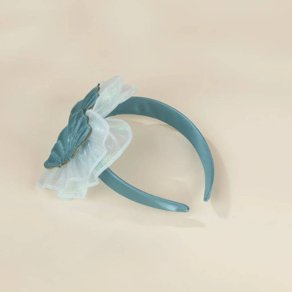 A teal blue headband featuring a decorative ruffled flower accessory in a dusty blue and white sheer fabric, displayed on a warm beige background.