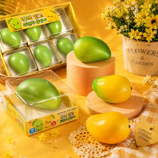 Green and yellow mango-shaped bath bombs displayed in clear plastic packaging with yellow cartoon labels, arranged on wooden blocks and a checkered cloth beside a basket of similar bath bombs and a flower pot on a yellow surface.