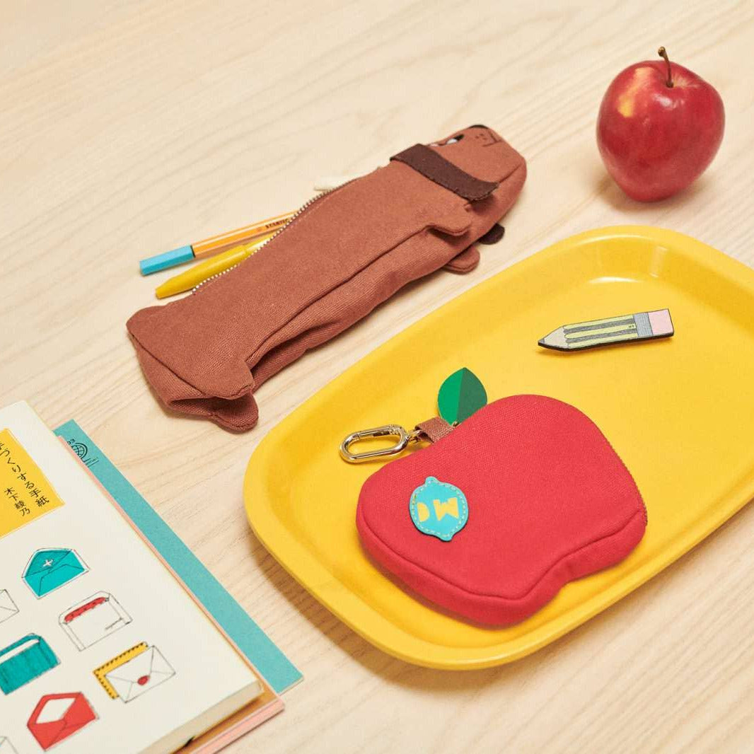 A school-themed felt accessory set arranged on a light wood surface, including a brown backpack-shaped pencil case, a red apple-shaped pouch with a blue apple sticker, a yellow tray, a pencil, a keychain, and an open notebook with colorful envelope illustrations. A red apple is also placed near the top right.