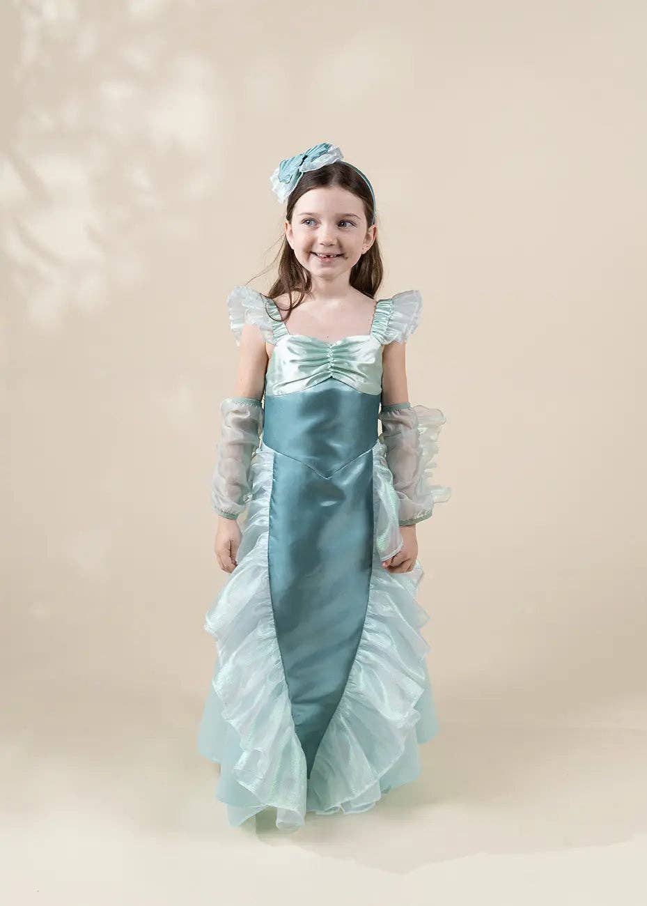 A young girl modeling a light blue mermaid-style costume dress with sheer ruffled sleeves, a satin bodice, and a matching headpiece against a soft beige studio background.