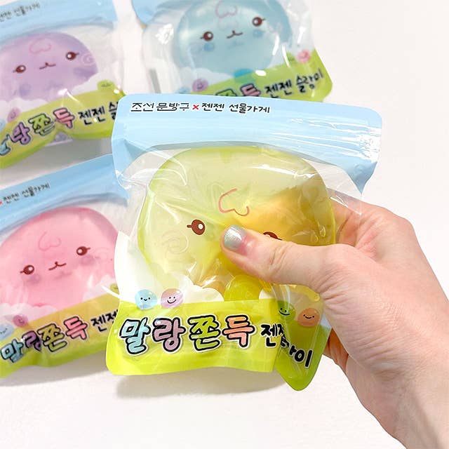 A hand holding a packaged yellow squishy toy in front of three more packaged squishy toys in pink, purple, and blue on a white background.