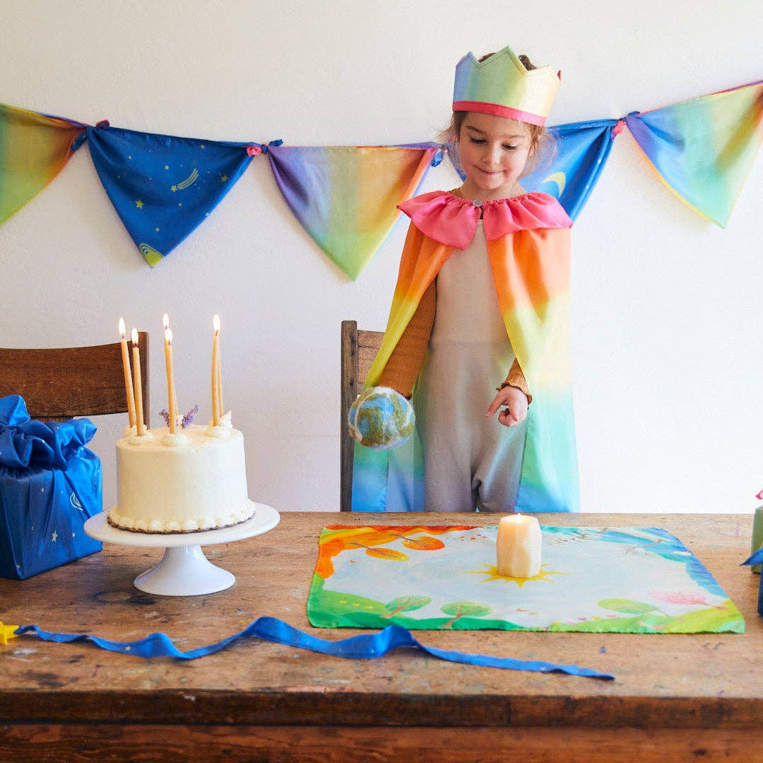Child in a rainbow-themed costume standing behind a table with a cake and decorations, against a colorful banner.