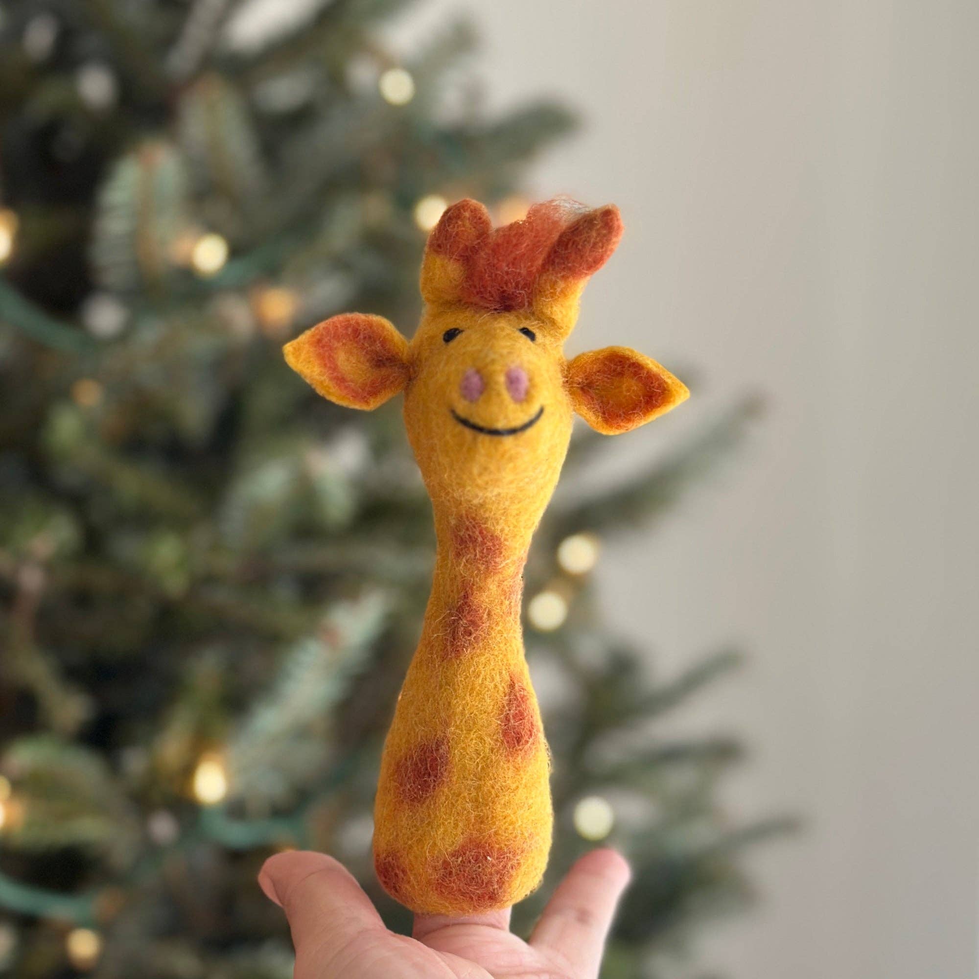 Hand holding a felt giraffe toy in front of a decorated Christmas tree.