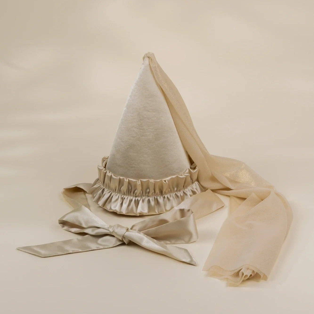 A cream-colored fabric pointed hat with a ruffled trim and long matching ribbon tails, photographed on a plain warm beige studio background.
