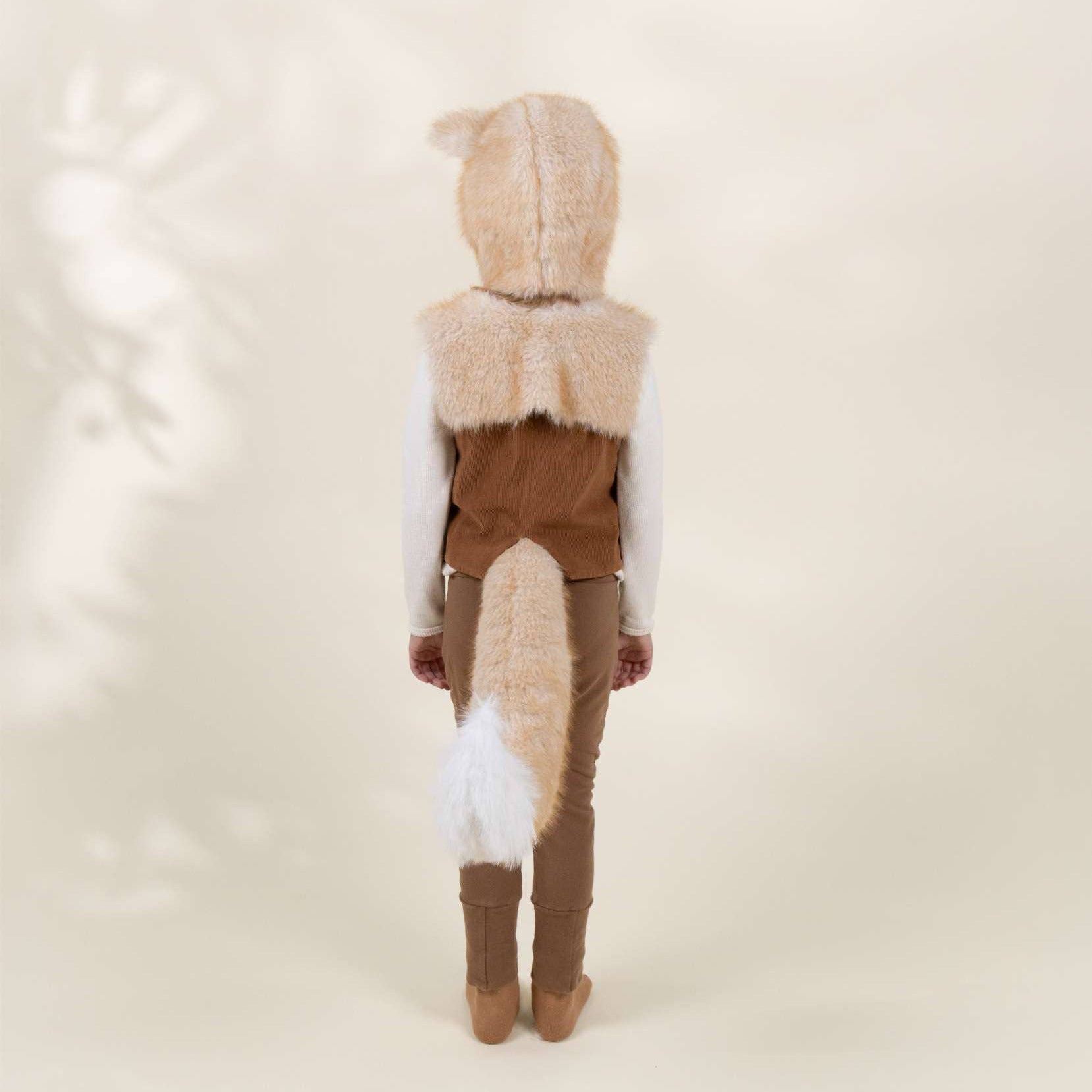 A child viewed from behind wearing a brown and beige animal costume with a faux-fur hood, vest, and tail, over a white long-sleeve shirt and brown pants, against a warm cream studio background.