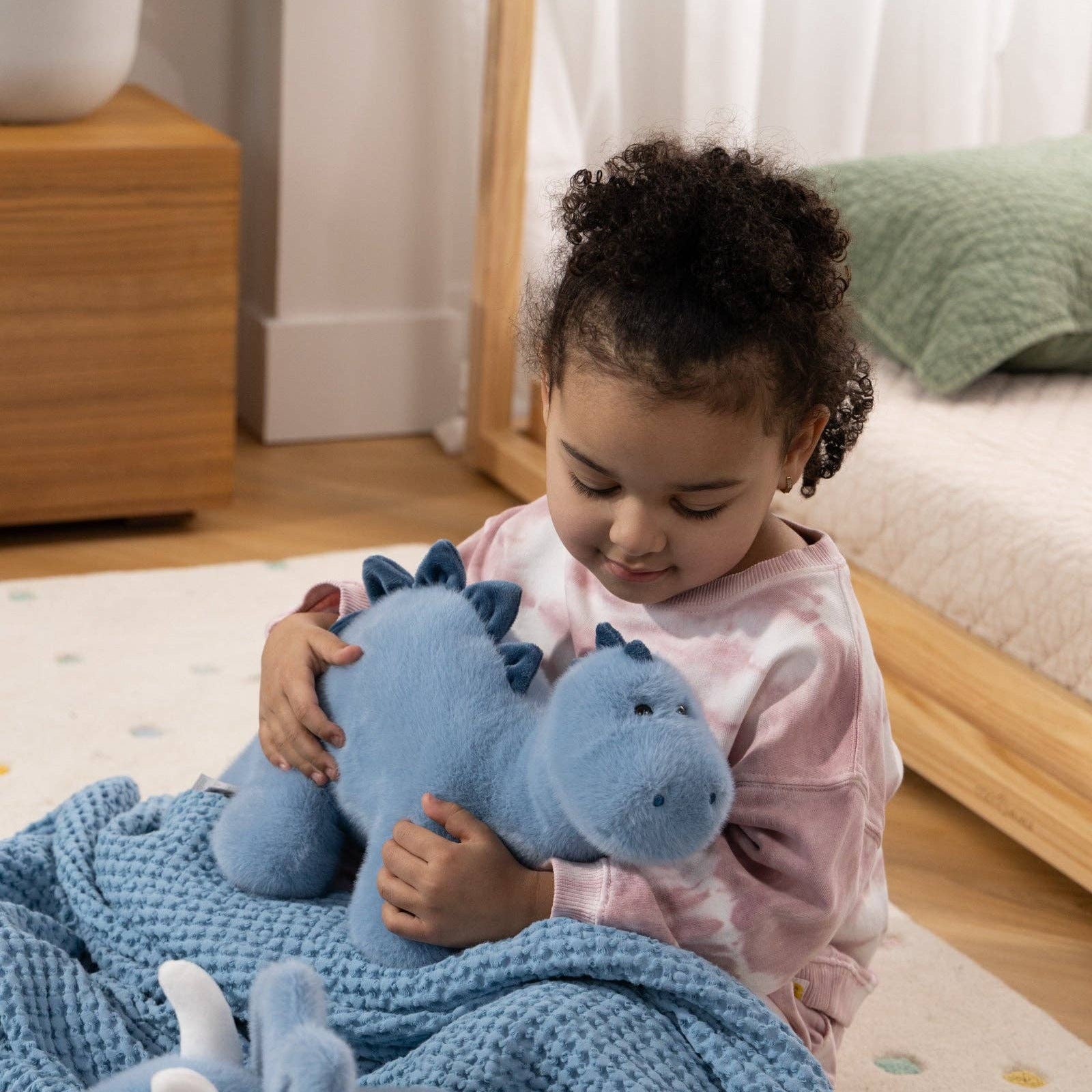 Child holding a blue dinosaur plush toy in a cozy room with a plant and wooden furniture.