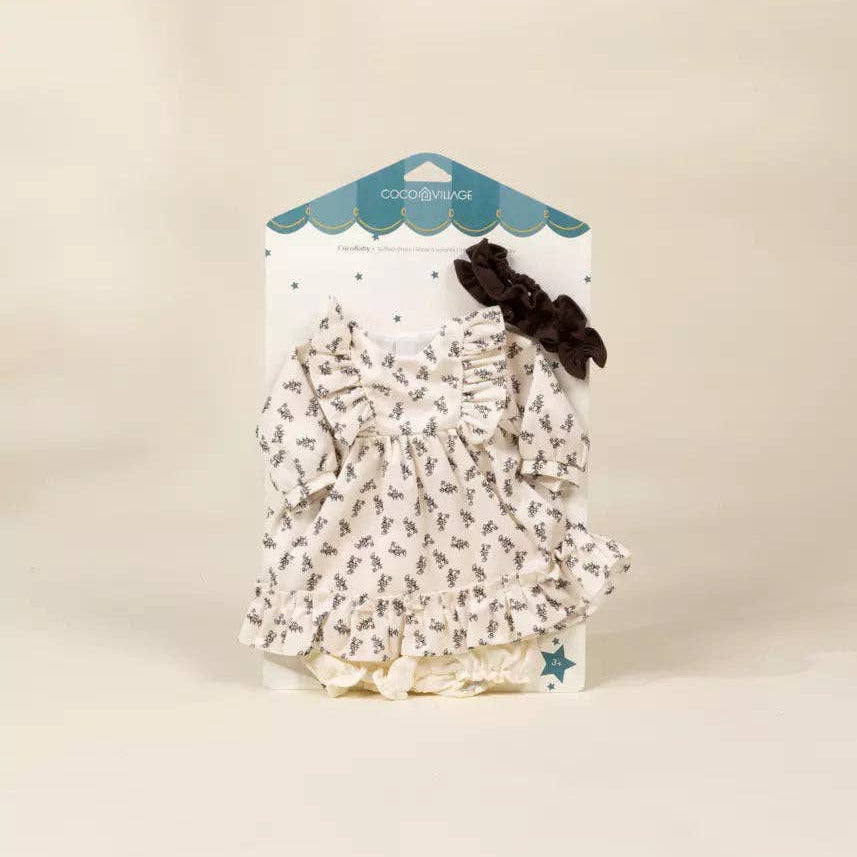 A doll dress set by Goodness Too, featuring a white ruffled dress with a brown floral pattern, a matching diaper cover, and a dark brown bow, displayed on its branded retail card against a warm beige background.