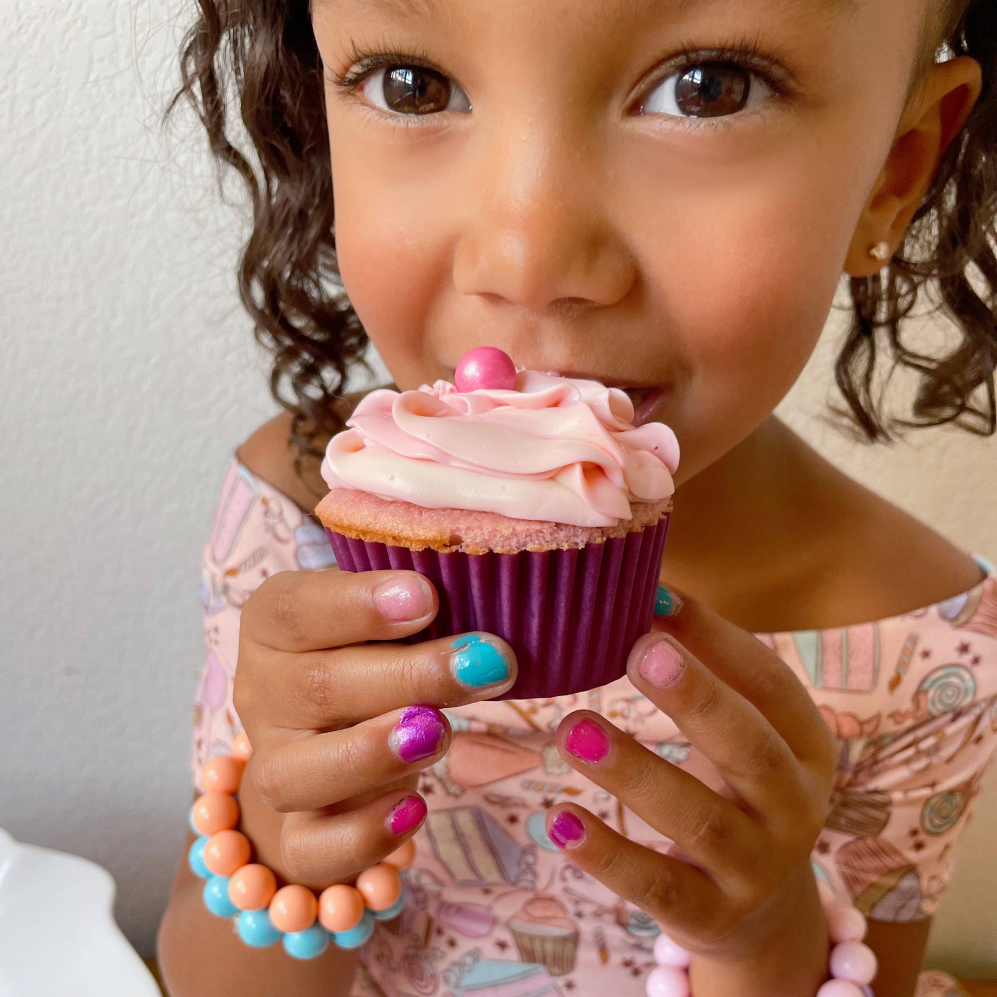 Child holding a pink cupcake with colorful frosting, wearing multiple bracelets.