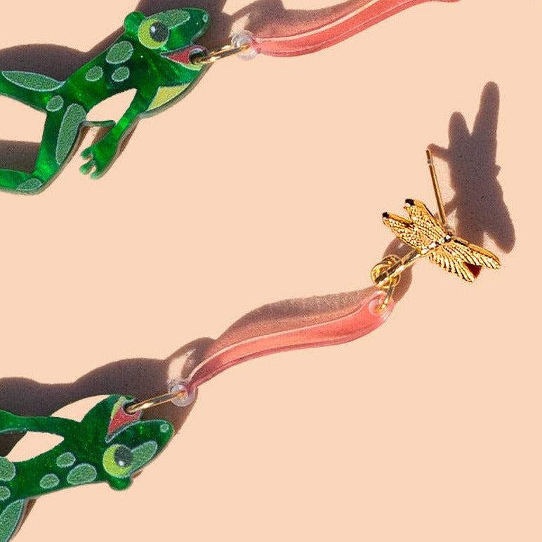 Frog and fly earrings with a pink tongue against a beige background