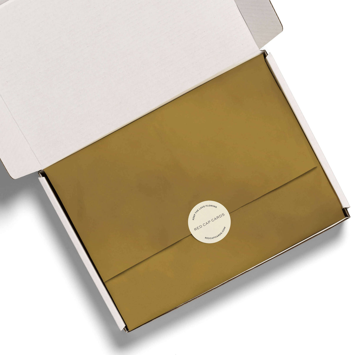 An open white presentation box containing a metallic gold envelope with a round white sticker seal that reads 'RED CAP CRAFTS'.