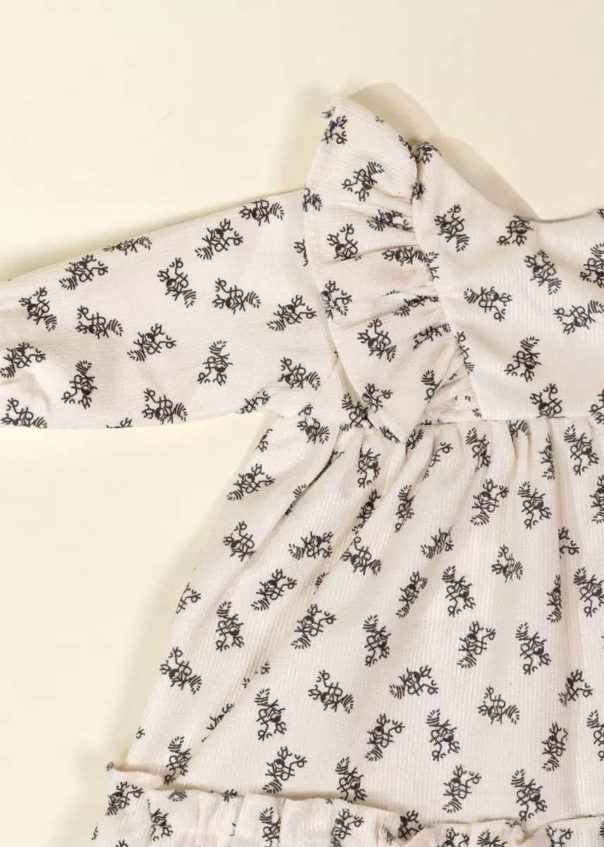 A close-up of a long-sleeve baby dress with a black floral block print on a warm cream-colored fabric, featuring a ruffled tie detail at the chest.