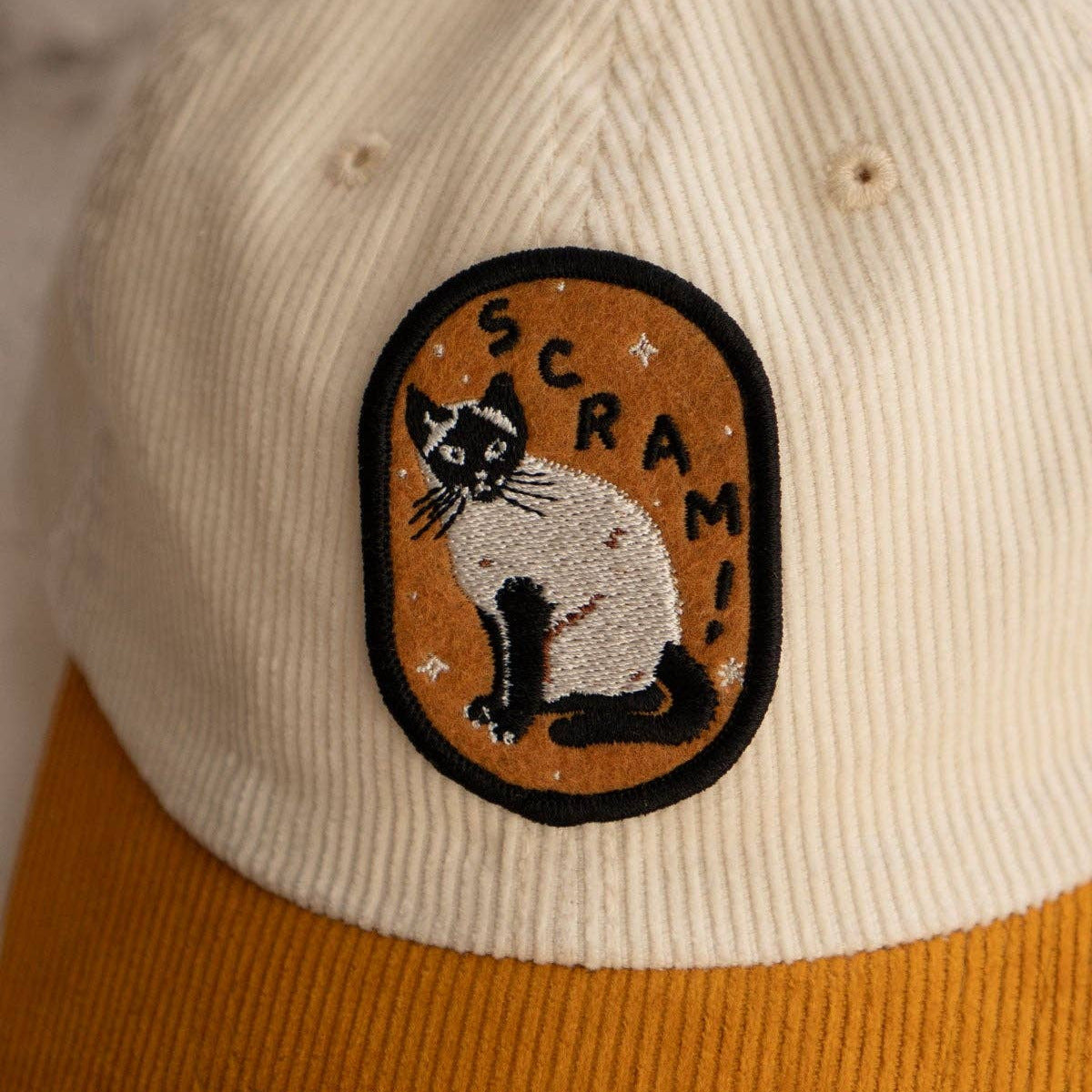 Beige and brown cap with a cat patch labeled 'SCRAM' on a blurred background