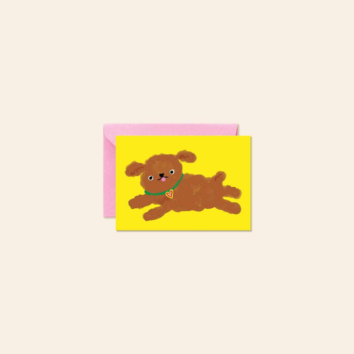 A greeting card featuring a hand-painted style brown puppy on a bright yellow background, shown with a pink envelope behind it on a light beige backdrop.