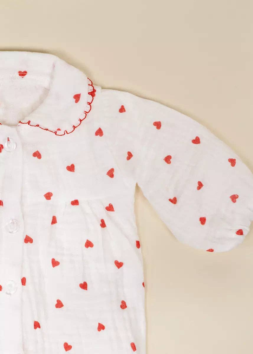 A close-up of a white long-sleeve baby garment, likely a muslin swaddle or sleeper, decorated with small red embroidered hearts and featuring a red scalloped collar.