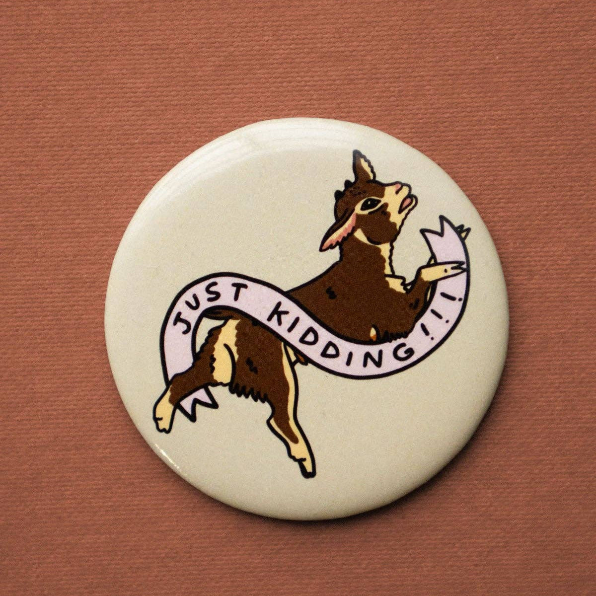 Button badge with cartoon goat holding a 'Just Kidding' sign on a brown background