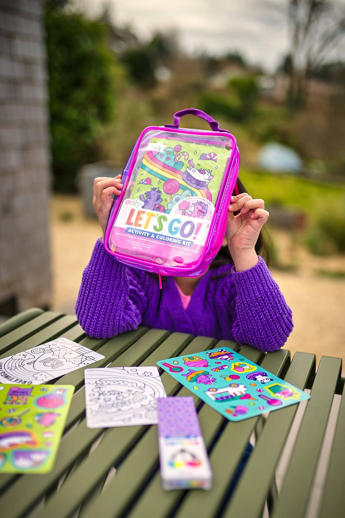 Child holding a colorful backpack with 'Let's Go!' text, surrounded by coloring books and stickers on a wooden table outdoors.