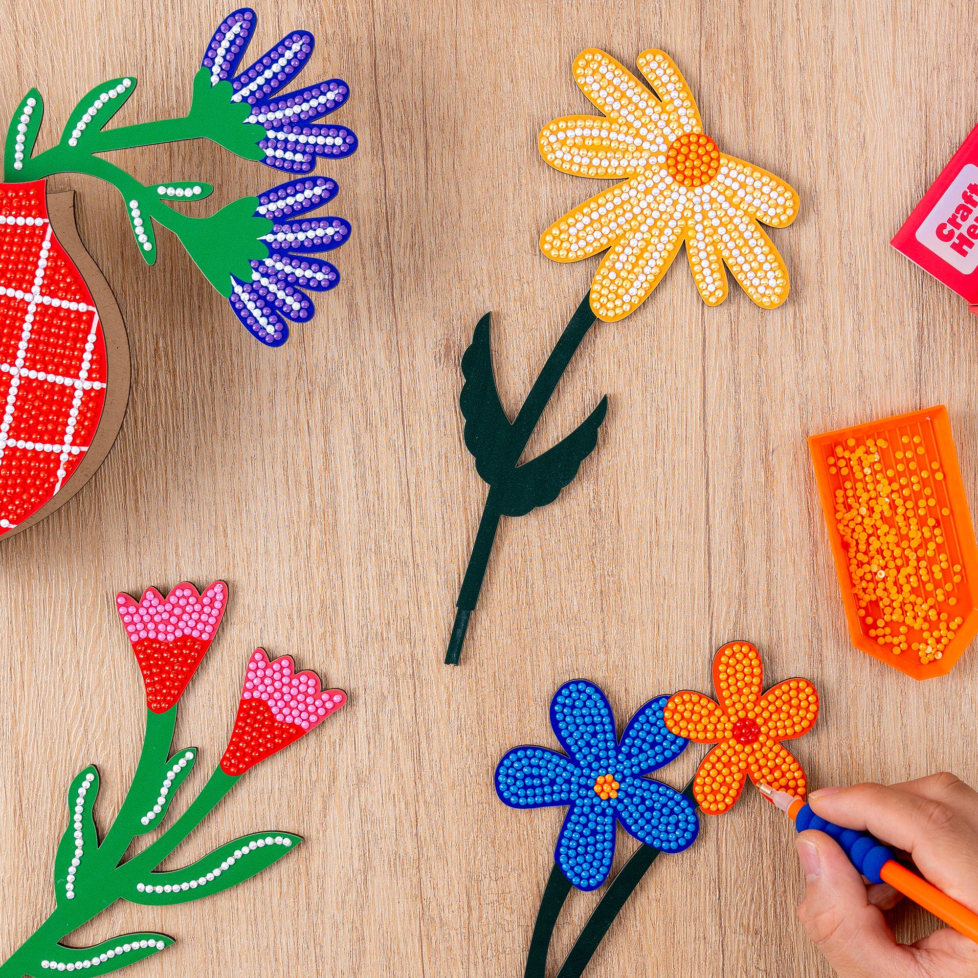 Colorful bead art flowers on a wooden surface with a hand using a tool.