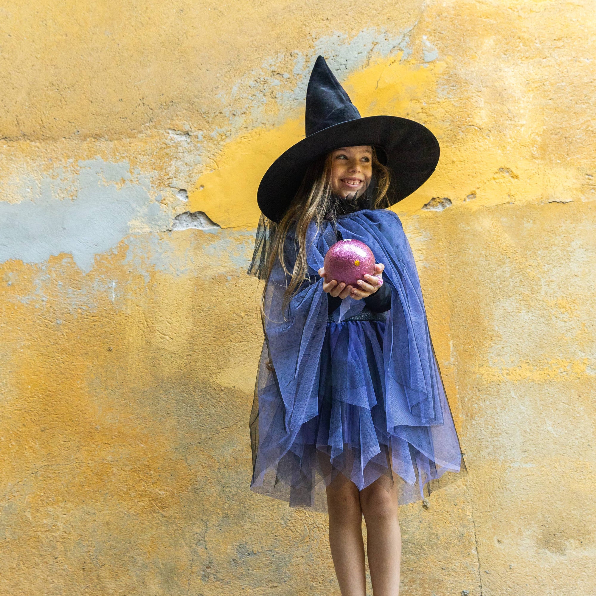 Child in witch costume holding a pink apple against a yellow wall.