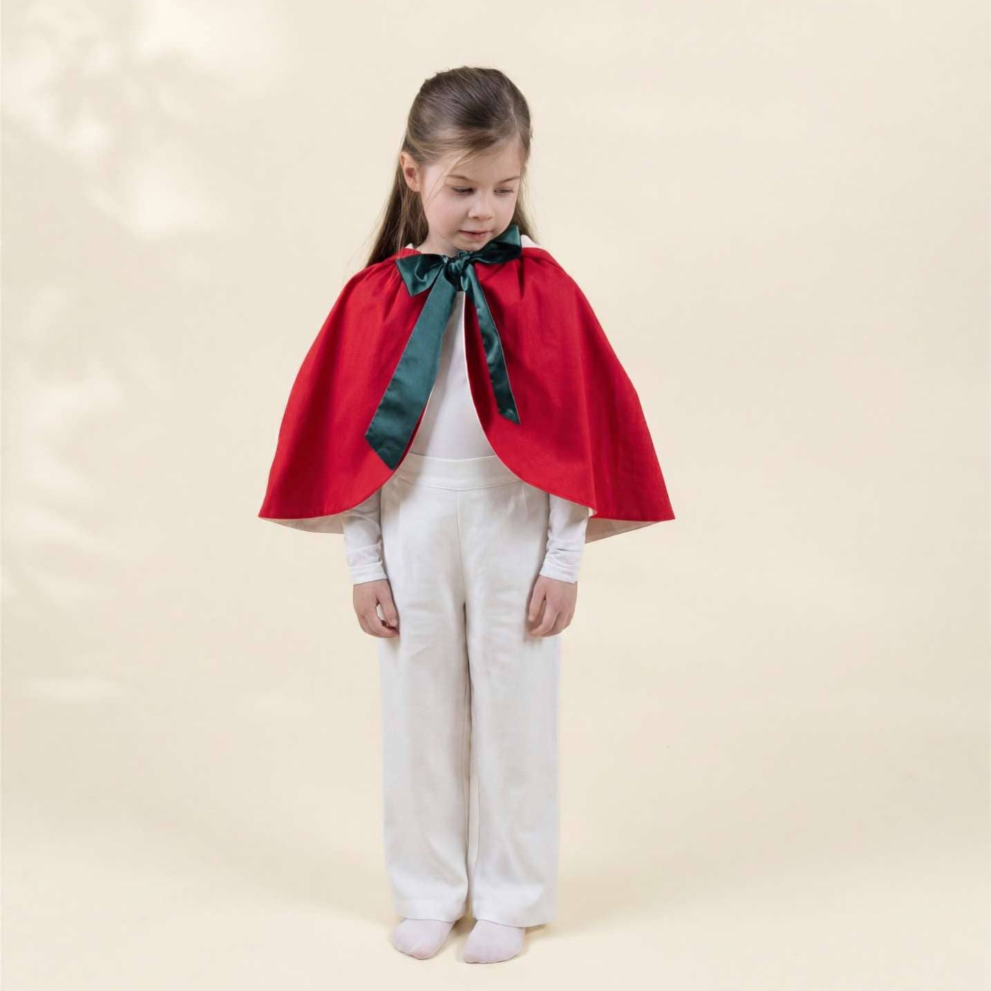 A young girl stands against a warm cream studio background wearing a bright red cape with a dark green satin neck bow over a white outfit.