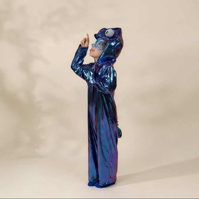 A child wearing a shiny holographic blue animal costume with a hood featuring a large eye detail, standing against a plain beige studio background.