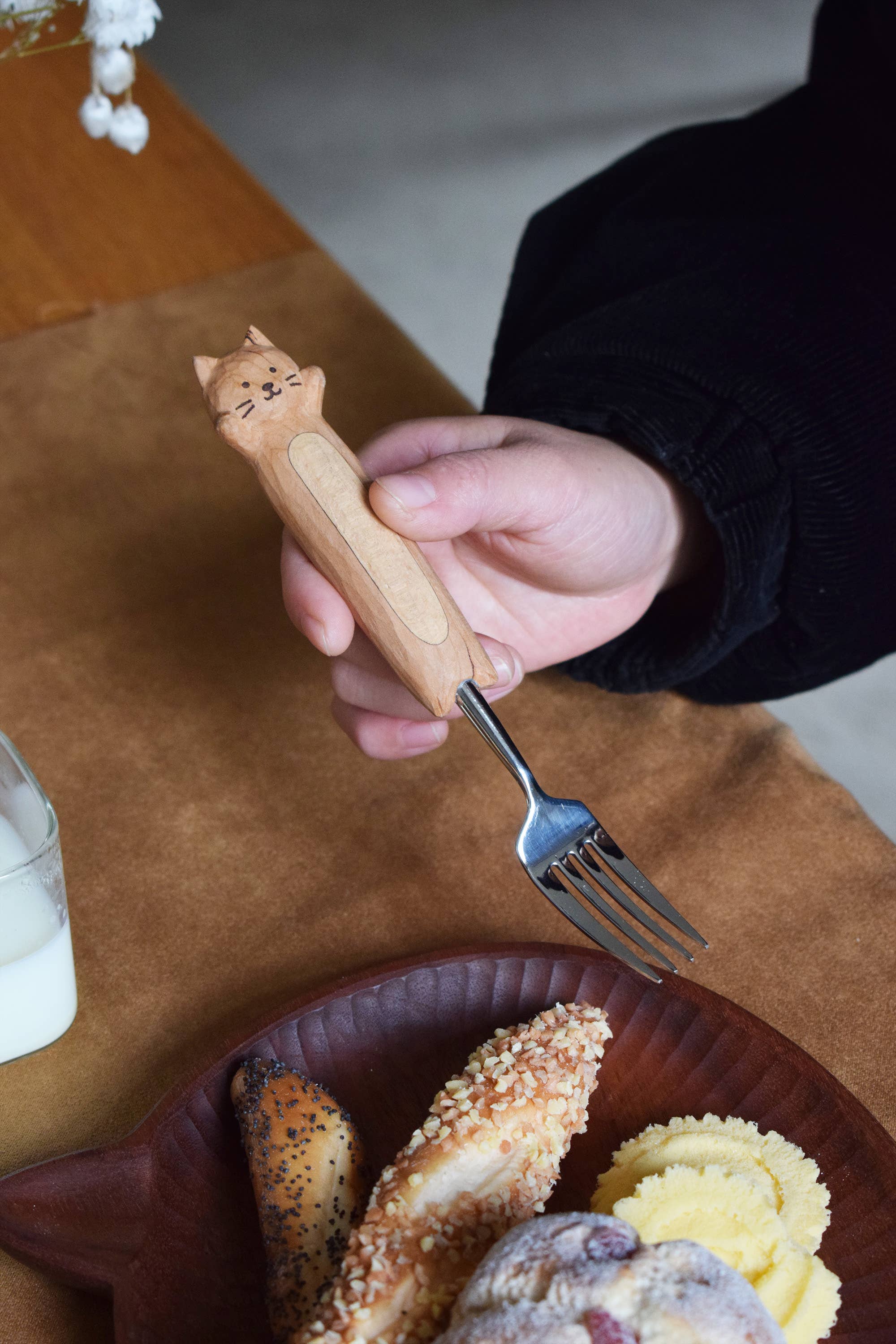 Hand holding a cat-shaped wooden fork over a plate of food on a wooden plate.